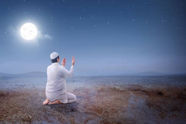 Rear view of asian muslim man sitting in pray position while raised hands and praying on the sand dune with moon and night scene background
